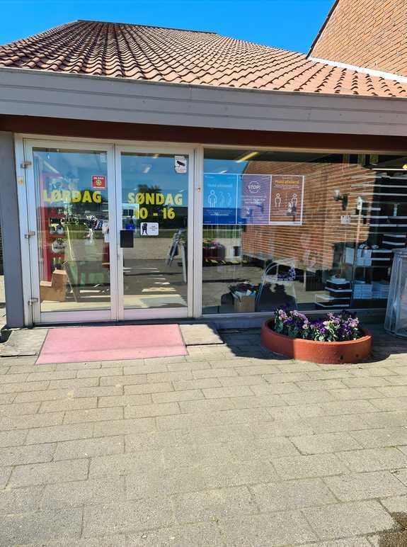 Butiksfacade med glasdøre, åbningstider og blomsterkummer foran. Solrigt vejr og blå himmel.