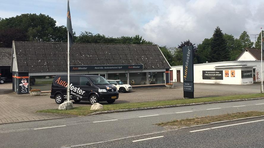 Bilforhandler med flere biler parkeret foran bygningen og en stor skilt ved vejen.
