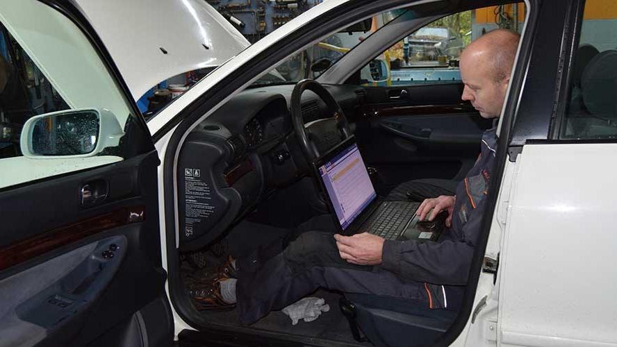 En mekaniker sidder i en hvid bil og arbejder på en bærbar computer i et værksted.