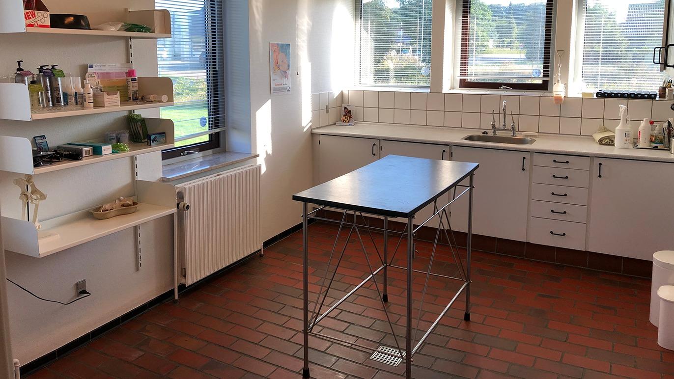 Køkken med bord, skabe og vinduer, der giver naturligt lys, i en lys og ren indretning.