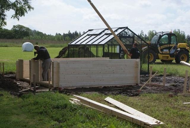 To bygge en træstruktur med håndværkere, en gartneri og en maskine i baggrunden på en græsmark.