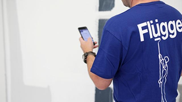 Mand i blå t-shirt med teksten "Flügger" står og bruger sin smartphone.