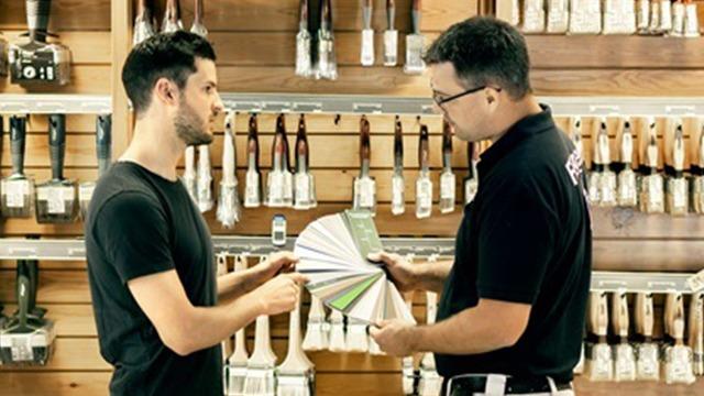 To mænd i en butik, der diskuterer farveprøver ved en væg med malerværktøj.
