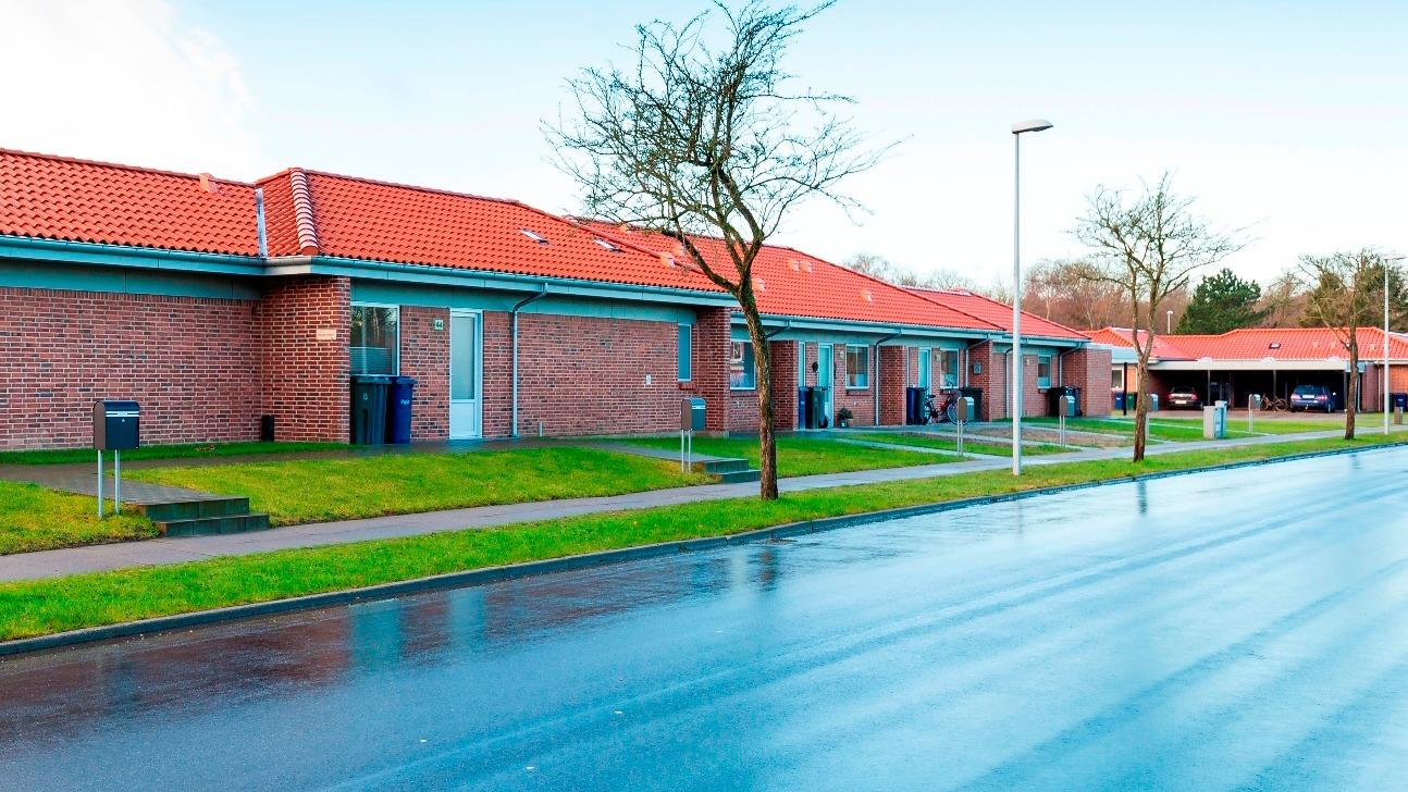 Rækkehuse med røde tage og græsplæne langs en våd vej i et boligområde.