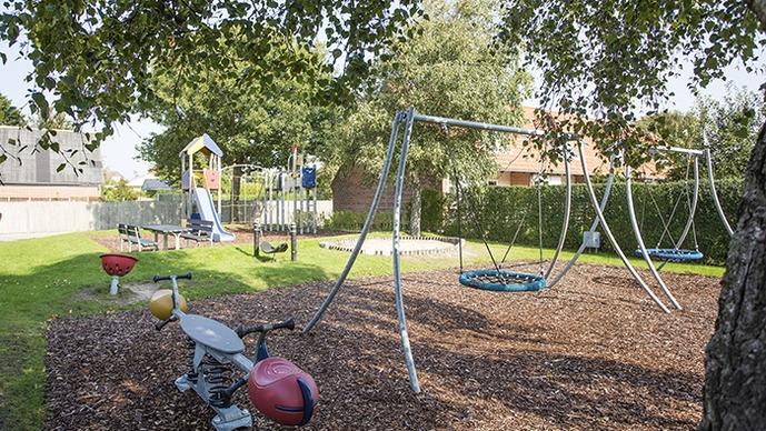 Legestativ med gynger og rutsjebane i en grøn park med træer og græs.