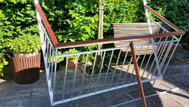 En metalramme med trædetaljer står i en have med grønne planter og en bænk i baggrunden.