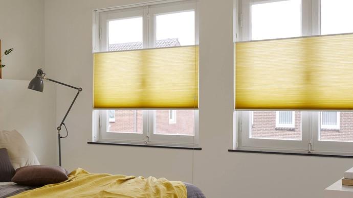 Soveværelse med gule plisségardiner i vinduerne og en seng med sengetøj i neutrale farver.