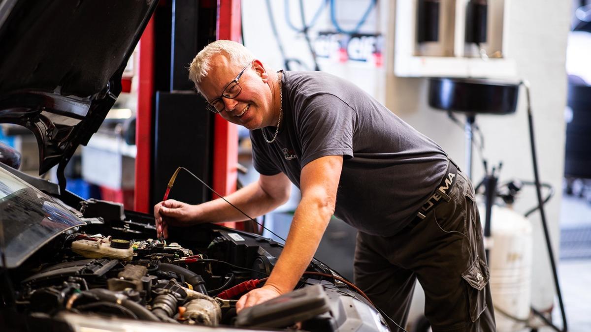 En mekaniker arbejder på en bilmotor i et værksted, mens han smiler og holder et værktøj i hånden.