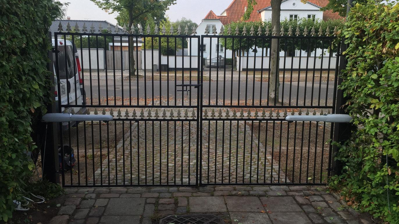 Sort jernport med to fløje, der åbner ind til en grusvej og en hvid murstensvilla i baggrunden.