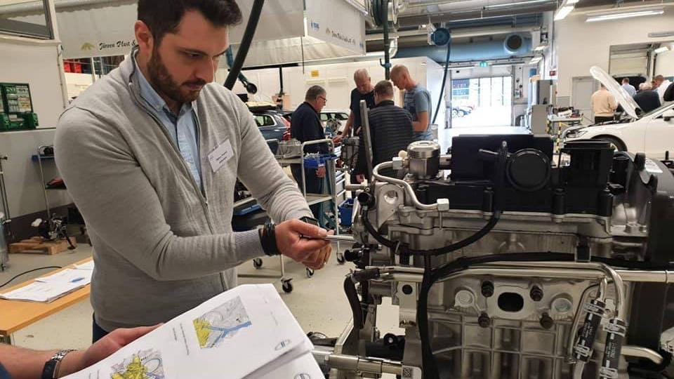 En mand arbejder med en motor, mens han ser på en manual i et værksted med andre personer i baggrunden.