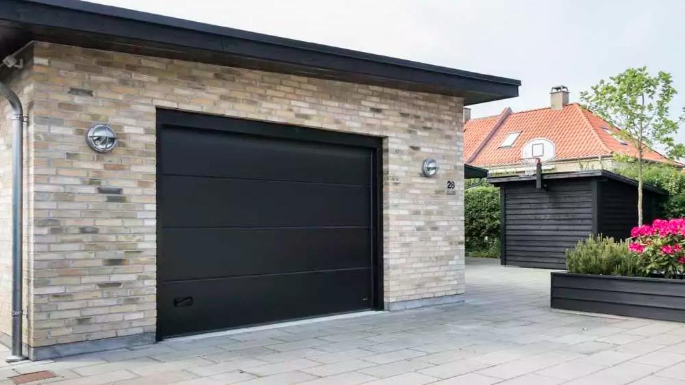 En moderne garage med sort garageport og murstensvæg, omgivet af grønne planter og blomster.