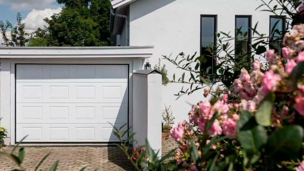 Hvid garageport ved en moderne hvid bygning omgivet af blomstrende buske og grønne træer.