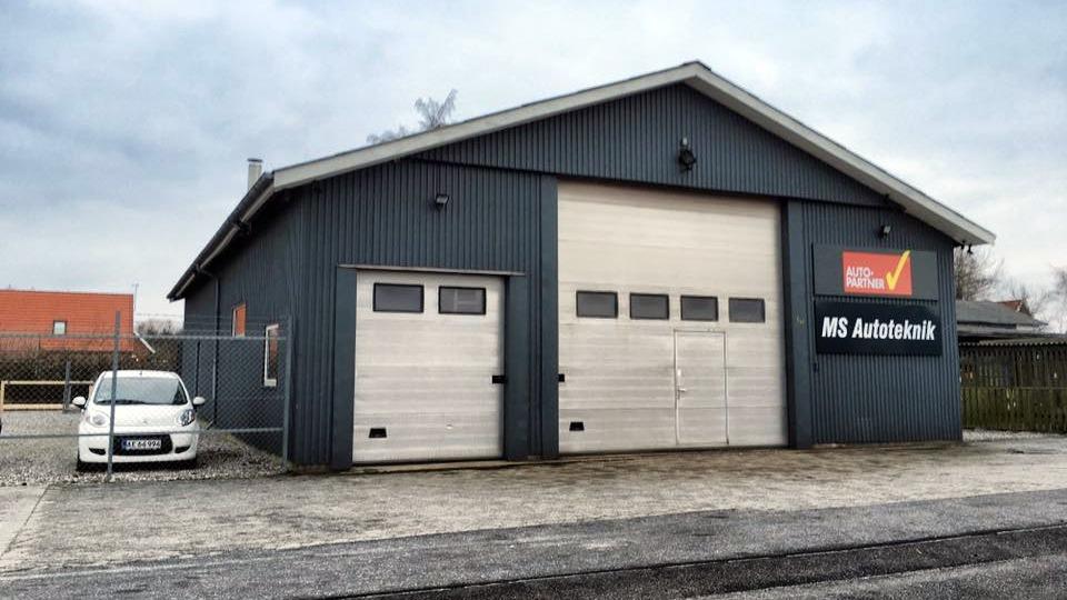 En grå autoværkstedbygning med to store porte og en hvid bil parkeret foran.