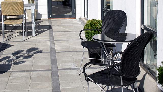 To terrasse med sorte stole og et lille bord, omgivet af fliser og grønne planter.