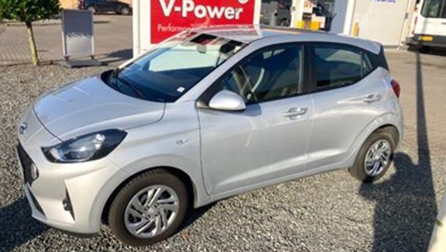 En sølvfarvet bil parkeret ved en tankstation med V-Power skilt i baggrunden.