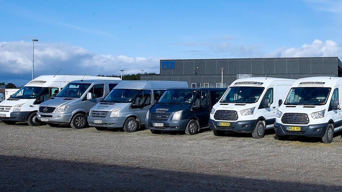 En række varevogne parkeret på en grusplads foran en bygning. Varevognene er i forskellige farver og modeller.