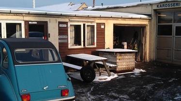 En blå vintage bil parkeret foran en bygning med sne på taget og en træbænk i gården.