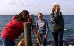 Tre personer står ved kysten, en kvinde kigger gennem kikkert, mens de andre ser ud mod havet.