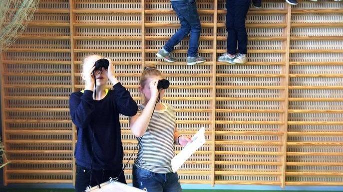 To personer står med kikkert og ser op, mens andre klatrer på en væg med træstiger.