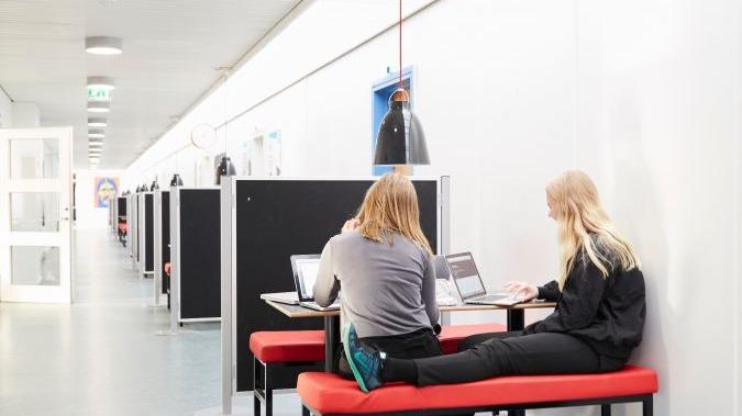 Studerende arbejder på bærbare computere ved et bord i en lys gang med arbejdsstationer.