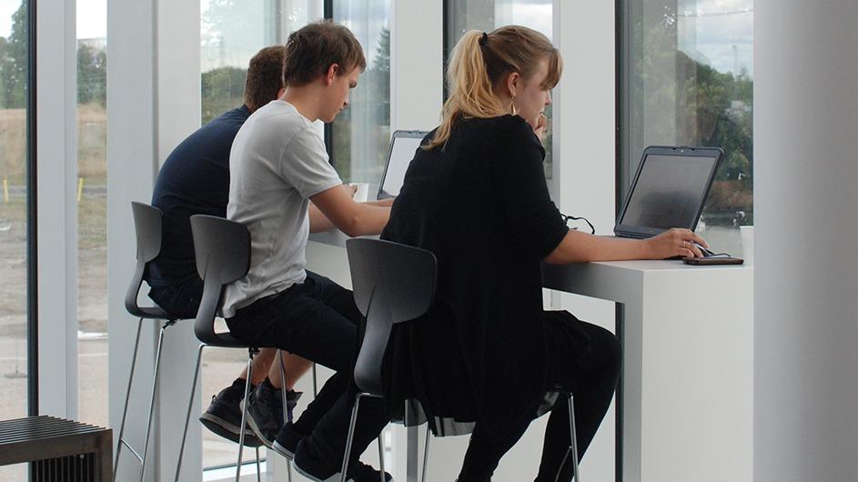 Tre unge mennesker sidder ved et bord og arbejder på bærbare computere i et lyst rum.