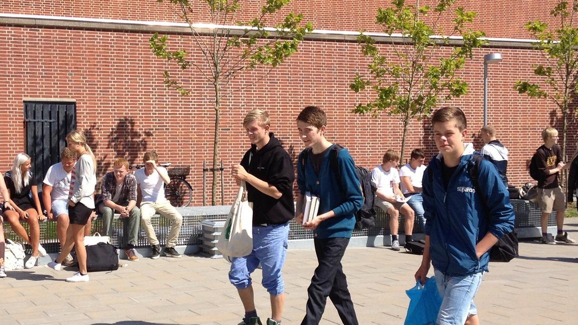 Tre unge drenge går på en brostensbelagt plads med træer og tilskuere i baggrunden.