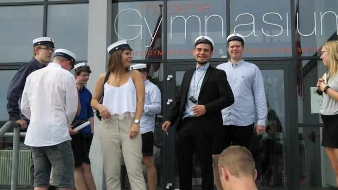 Gruppe af studerende i studenterhuer, der fejrer dimission foran gymnasiet. Solrigt vejr og glade ansigtstræk.