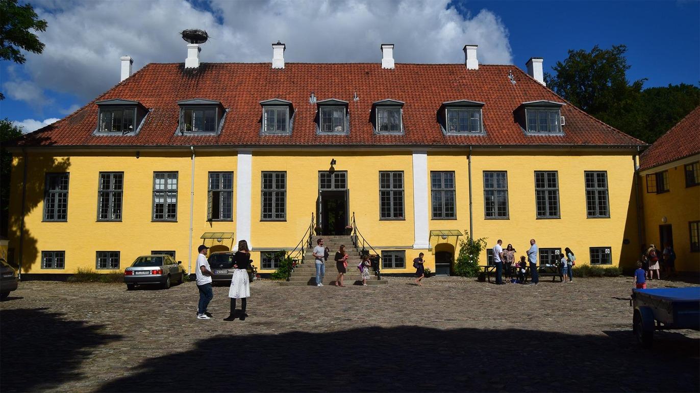 Gul bygning med mange vinduer og mennesker foran, omgivet af grønne træer og blå himmel.