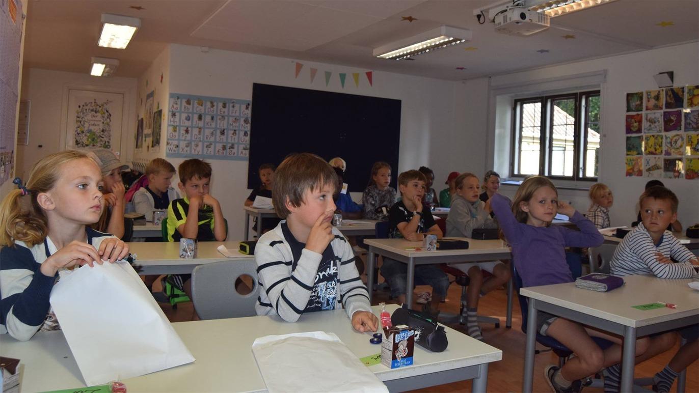 Børn sidder i et klasseværelse, opmærksomme og engagerede i undervisningen ved deres borde.
