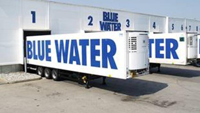 Tre lastbiler med store blå bogstaver, der står "BLUE WATER", parkeret foran lagerbygninger.