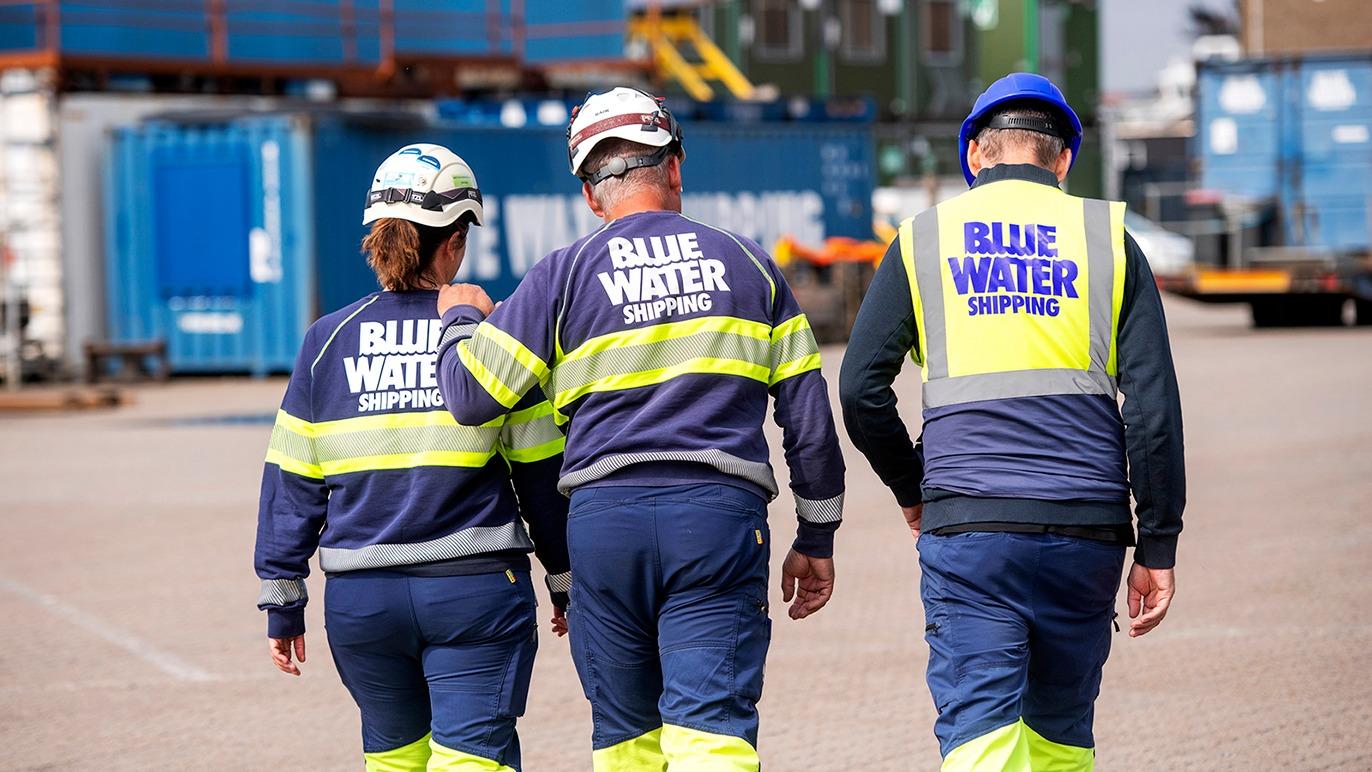 Tre arbejdere fra Blue Water Shipping går sammen på en havn med containere i baggrunden.