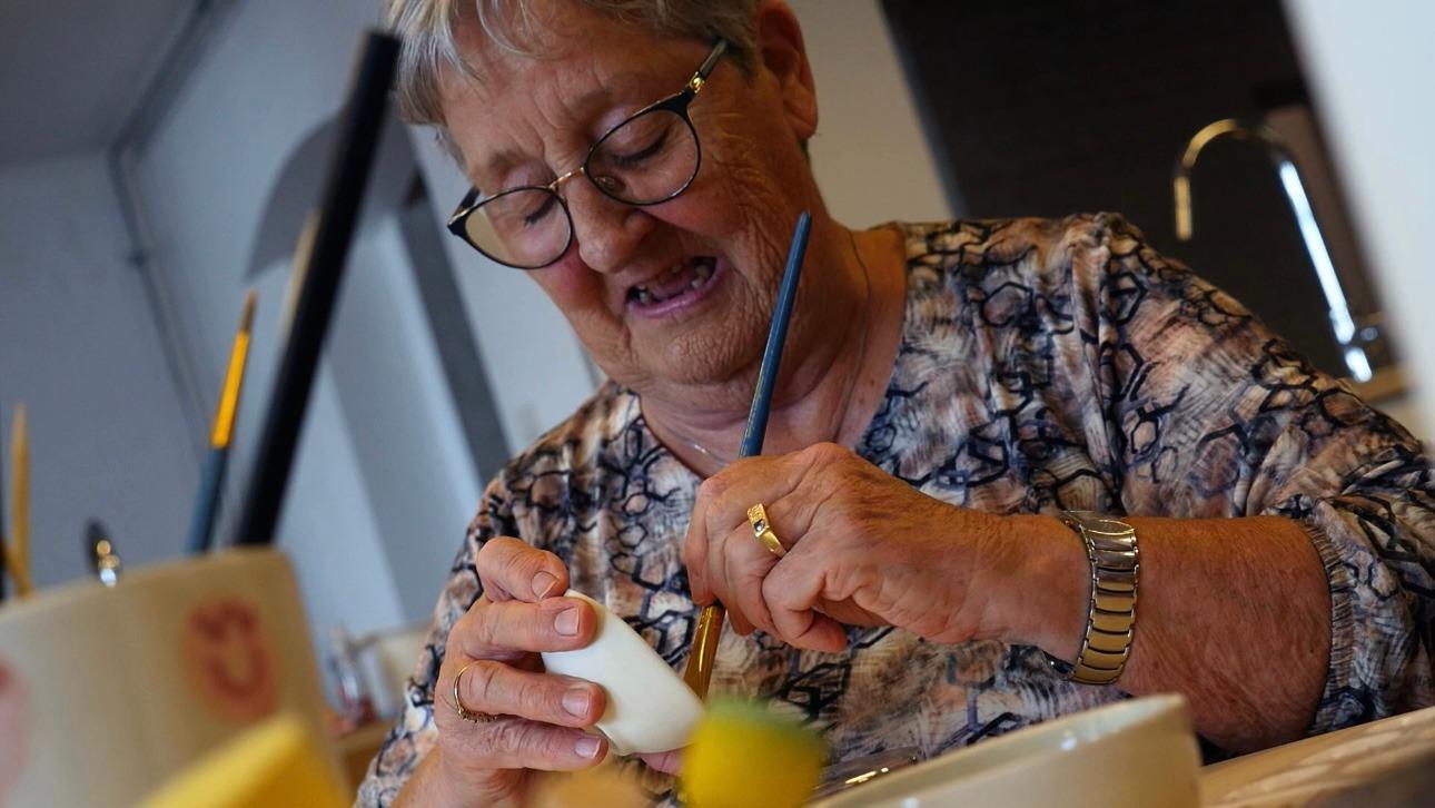 Ældre kvinde maler på et æg med pensel, omgivet af farverige dekorationer og værktøjer i et lyst rum.