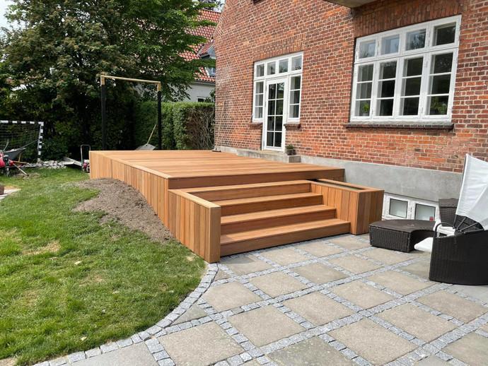 Terrasse i træ med trin, græsplæne og flisebelægning ved en murstensvæg og vinduer.