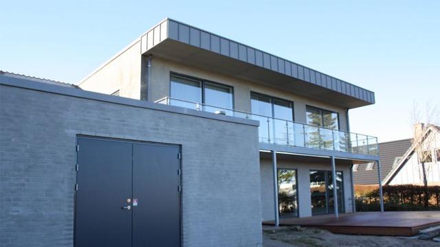 Moderne hus med stor balkon og grå facade, beliggende i et roligt boligområde.