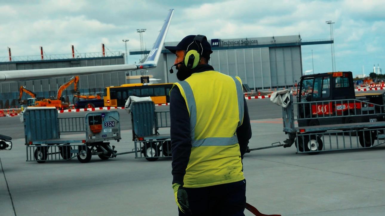En lufthavnsarbejder iført sikkerhedsvest og høreværn står ved flyet på en travl lufthavn.