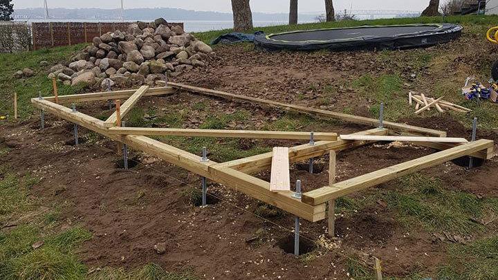 Byggeri af en træramme på fundamentpæle i en have med græs og sten.