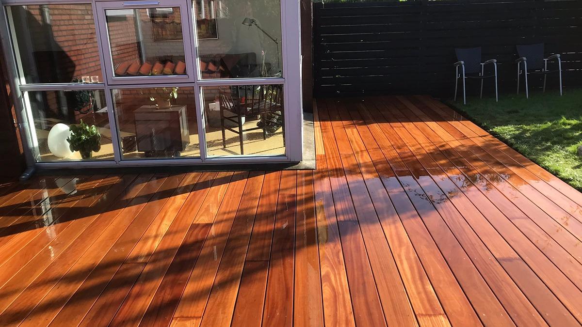 En solbeskinnet terrasse med nybehandlede træplanker og et vindue, der viser indendørs møbler.