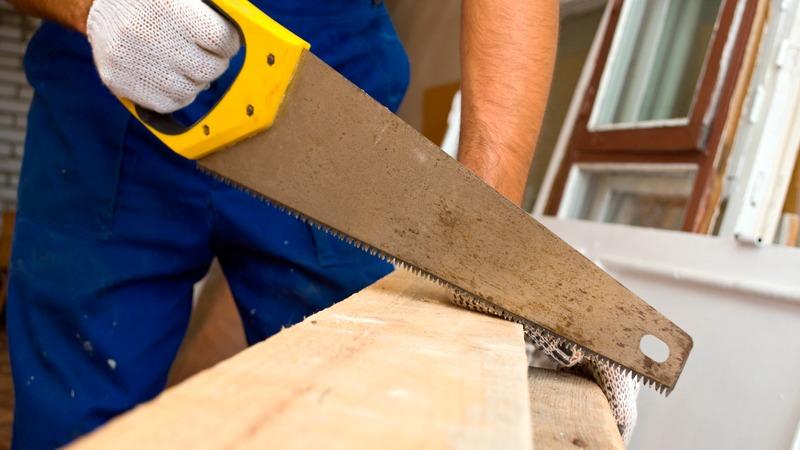 En håndværker saver i et stykke træ med en sav, iført handsker og blå arbejdstøj.
