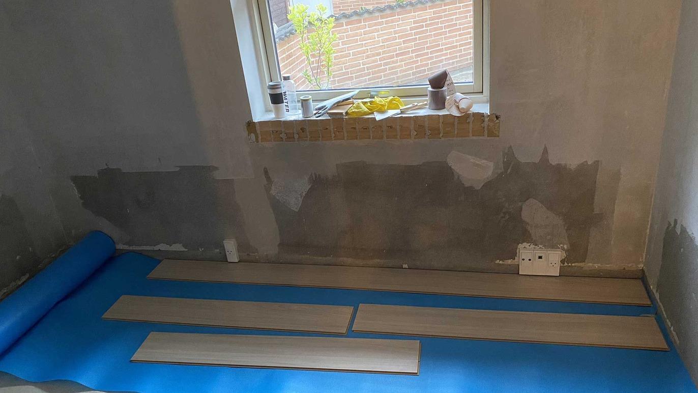 Gulvbelægning i en stue med blå underlag og træplanker klar til installation ved vinduet.