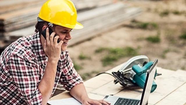 En håndværker i gul hjelm taler i telefon og arbejder på en bærbar computer ved en byggeplads.
