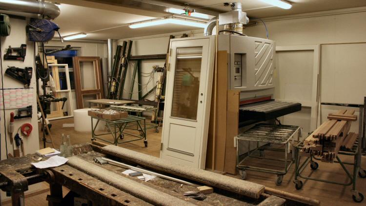 Et værksted med en stor maskine, træplader og værktøj på arbejdsbænke, der viser en travl arbejdsplads.