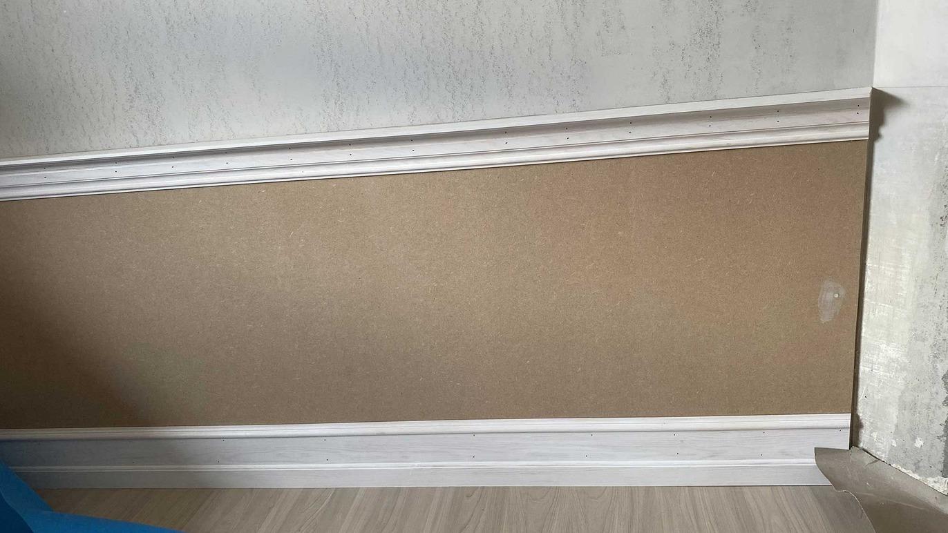 Væg med beige tapet og hvid fodliste i et rum med lys trægulv.
