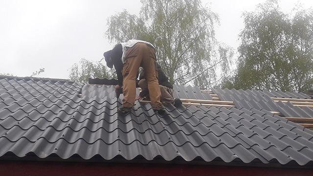 En håndværker arbejder på et tag med metalplader, mens han står på en stige under en grå himmel.