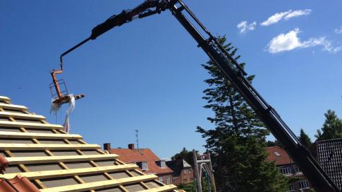 Kran løfter en stige op til et tag under en klar blå himmel med få skyer.
