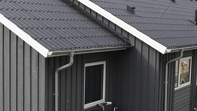 Taget af et moderne hus med sort metaltag og grå træfacade, der viser vinduer og tagrender.