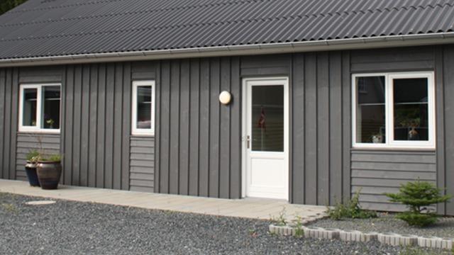 Moderne grå træhus med hvide vinduer og dør, beliggende på en grusvej.