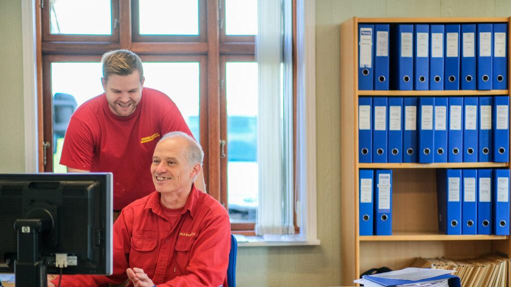 To mænd i røde skjorter arbejder sammen ved en computer i et kontor med blå ringbind i baggrunden.