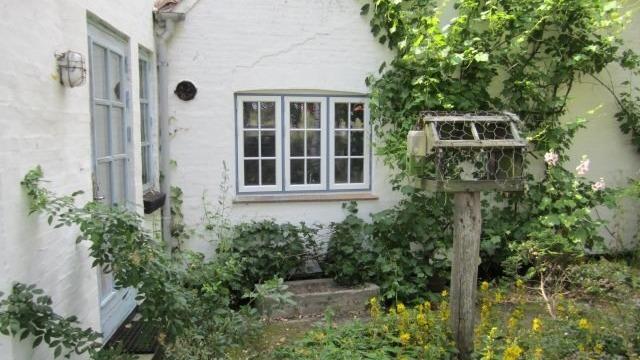 Hvidt hus med vinduer, omkranset af grønne planter og en fuglefoderstation i haven.