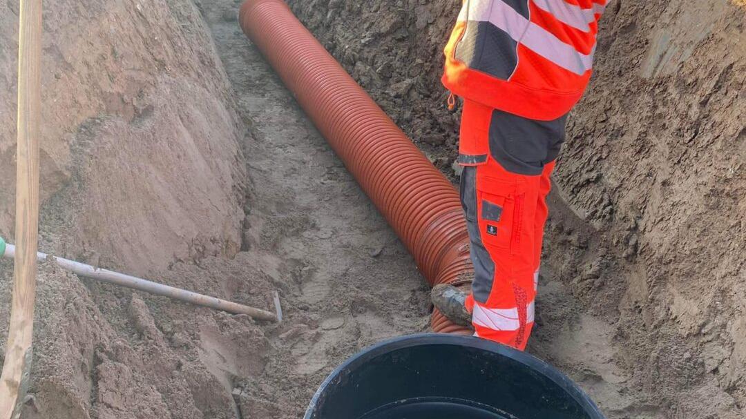 Arbejder i en grav med orange rør og en sort spand, iført sikkerhedstøj og handsker.