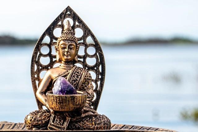 Buddha-statue med ametyst i skålen, placeret ved vandet med en rolig baggrund.